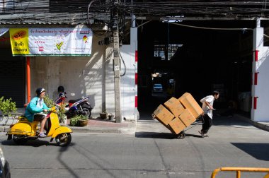 Bangkok, Tayland - Aralık 2015: Sokak pazarında karton kutularla el arabası taşıyan adam. Yolda sarı motosikletini süren başka bir adam..