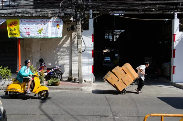 Bangkok, Tayland - Aralık 2015: Sokak pazarında karton kutularla el arabası taşıyan adam. Yolda sarı motosikletini süren başka bir adam..