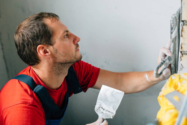 Man in red shirt and blue overall with putty knife doing putty