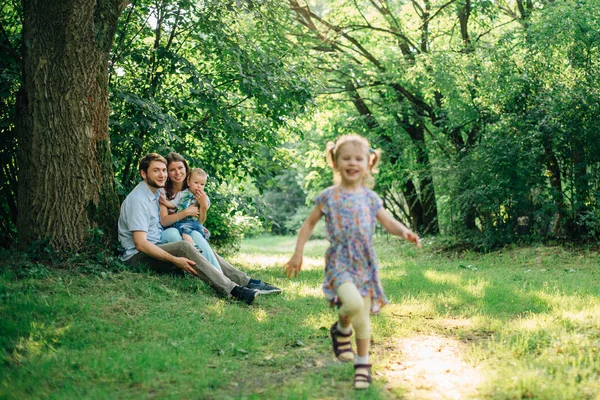 Young happy family playing in the park. Little smiling girl running from her parents and brother. Man sitting with his wife and son looking at his cute daughter.