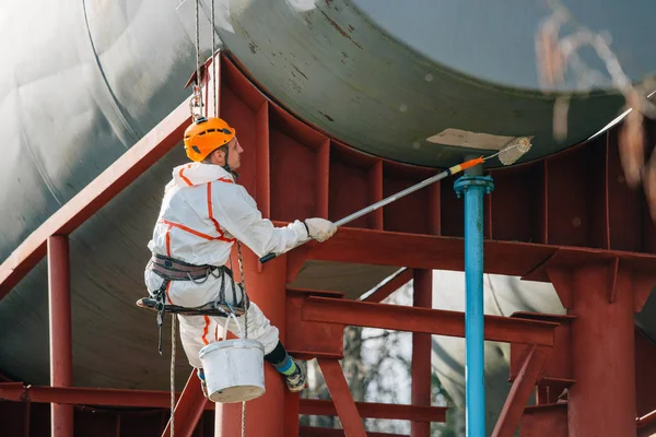 Industrial climber in helmet and uniform painting water tower. Professional Painter working on height. Risky job. Extreme occupation.