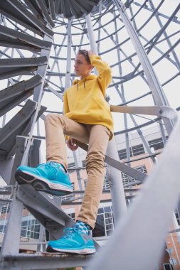 Sarı kapüşonlu ve turkuaz spor ayakkabılı genç bir adam spiral bir merdivende duruyor. Genç güzel mimaride güzel bir spiral merdivenle atlıyor.
