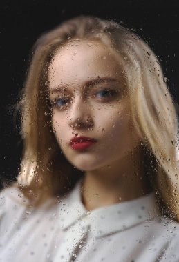 Beyaz gömlekli güzel bir kız su damlalarıyla dolu bir bardağın arkasında üzgün. Kör bir bardağın arkasında genç ve narin bir kadının fotoğrafı.