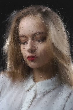 Beyaz gömlekli güzel bir kız su damlalarıyla dolu bir bardağın arkasında üzgün. Kör bir bardağın arkasında genç ve narin bir kadının fotoğrafı.