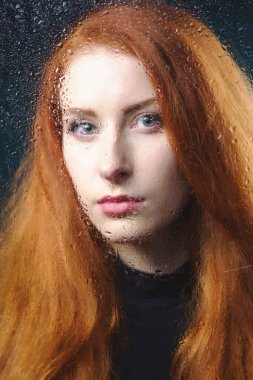 Siyah balıkçı yakalı güzel, kızıl saçlı bir kız su damlalarıyla dolu bir bardağın arkasında üzgün. Kör bir camın arkasında genç, çekici bir kadının fotoğrafı.