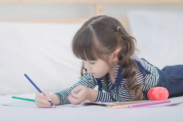 Aranyos ázsiai kislány csinál házi feladatot, írás, színes ceruzák ágyban otthon. — Stock Fotó