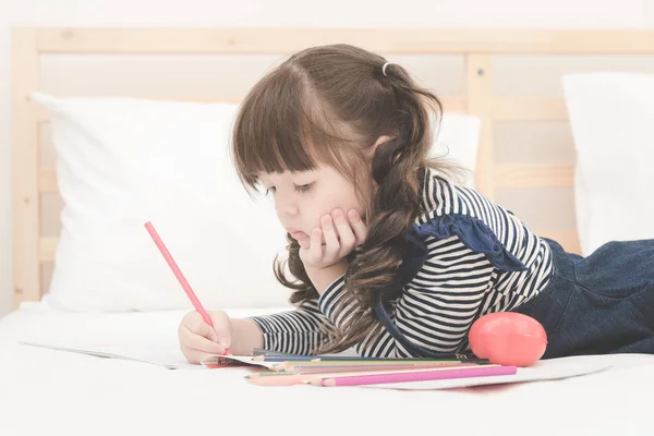 Linda niña asiática haciendo la tarea, escribiendo con lápices de colores en la cama en casa . —  Fotos de Stock