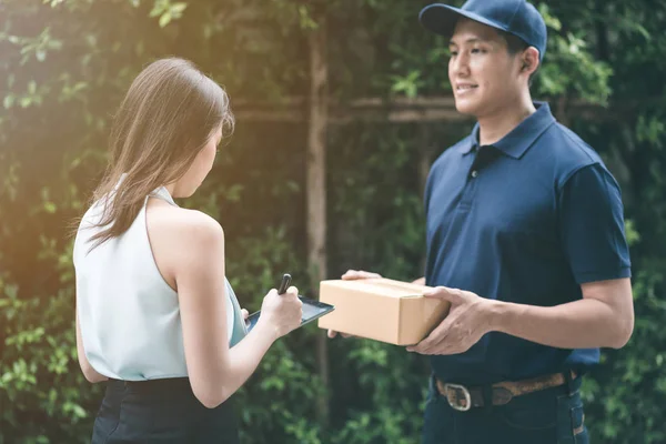 Guapo joven asiático entrega hombre sonriendo y sosteniendo una caja de cartón mientras espera hermosa joven mujer asiática poner firma en tableta inteligente para confirmar recibir . — Foto de Stock