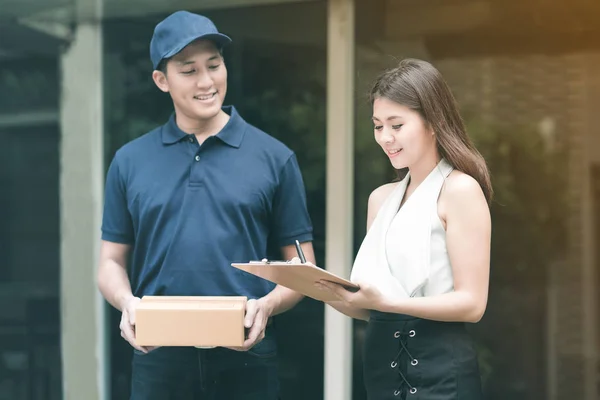 Knappe jonge Aziatische levering mens glimlachend en houden van een kartonnen doos terwijl u wacht mooie jonge Aziatische vrouw handtekening aanbrengend Klembord voor bevestigen ontvangen. — Stockfoto
