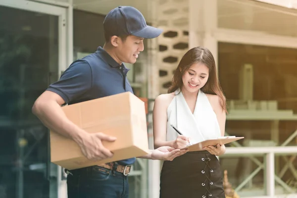 Knappe jonge Aziatische levering mens glimlachend en houden van een kartonnen doos terwijl u wacht mooie jonge Aziatische vrouw handtekening aanbrengend Klembord voor bevestigen ontvangen. — Stockfoto