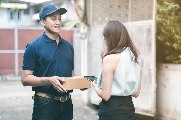 Handsome young asian delivery man smiling and holding a cardboard box while waiting beautiful young asian woman putting signature in smart tablet for confirm receive. — Stock Photo, Image