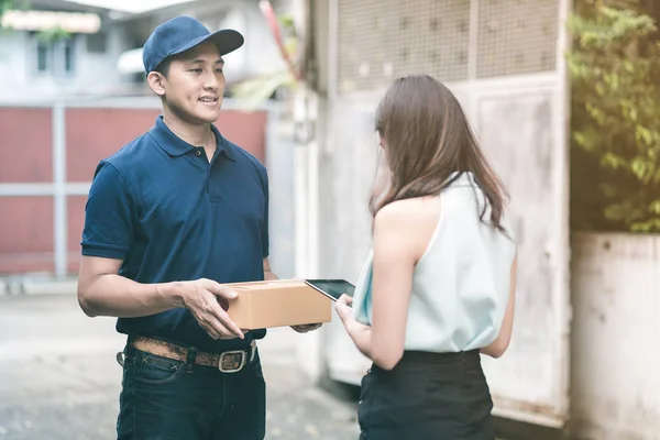 Guapo joven asiático entrega hombre sonriendo y sosteniendo una caja de cartón mientras espera hermosa joven mujer asiática poner firma en tableta inteligente para confirmar recibir . — Foto de Stock