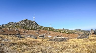 Rüzgâr türbini, bitki örtüsü, granit taşlar ve dört çeker aracın olay yerinde hareket ettiği bir dağ manzarası