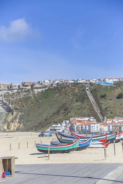 Typowy widok na plażę Nazare z kolorowych i tradycyjnych łodzi rybackich i klifu z kolejki linowej i miasta na szczycie jako tło, w Portugalii — Zdjęcie stockowe