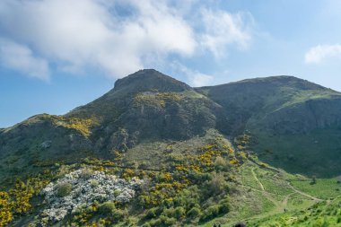 Holyrood Park 'taki yeşil dağa bakın. İnsanlar arka planda yürüyüş, bitki örtüsü ve mavi bulutlu gökyüzüne bakıyor.
