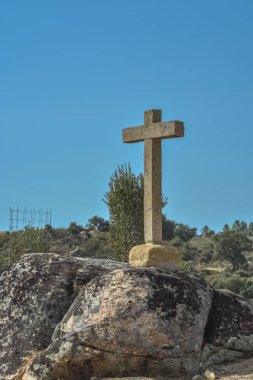 Taştan haç heykelinin detaylı görüntüsü, mavi gökyüzü ve arka plandaki ağaçların tarlası