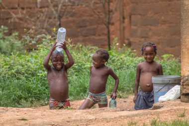 Sumbe / Angola - 02 25 2020: Biri plastik su şişesiyle banyo yapan üç Afrikalı çocuğun görüntüsü, diğeri ona bakıyor ve kız etkileyici bir görünüm sergiliyor