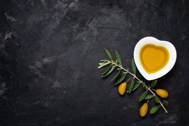 Durgun hayatın üst görüntüsü. Seramik kasede, kalp şeklinde, ekstra saf zeytinyağı, kaba siyah arka planda yeşil zeytinli bir dal. Metin için boş alan