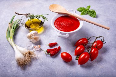 Kırsal bir arka planda, domates soslu beyaz bir seramik kase, fesleğen yaprağı, küme domates, zeytinyağı ve çeşitli baharatlar.