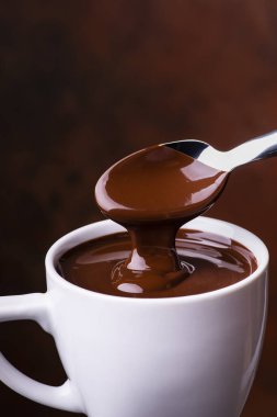 Ön planda, sıcak ve sıvı çikolata kaşıktan beyaz bir seramik kaba dökülüyor.