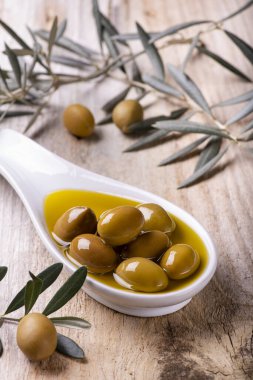 Ön planda, beyaz bir seramik kase, ekstra saf zeytinyağı ve yeşil zeytinyağı, kırsal ahşap masa üzerinde zeytin dalı