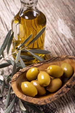 Ön planda, kırsal ahşap masada, yeşil zeytinli ahşap bir kase, zeytin dalı ve ekstra saf zeytinyağı.