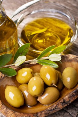 Ön planda, kırsal ahşap masada, yeşil zeytinli ahşap bir kase, zeytin dalı ve ekstra saf zeytinyağı.