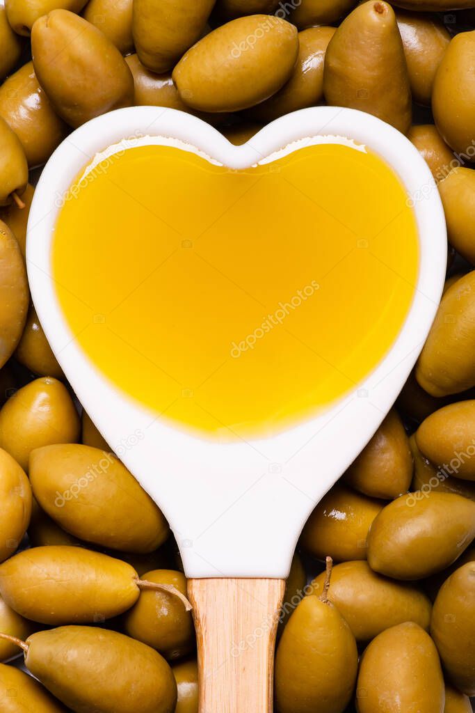 In the foreground, with a view from above, background with green olives and white spoon, heart-shaped, with extra virgin olive oil.