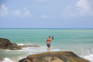 Kayanın üzerinde duran, bir kamera önünde dalga elinde tutarak turist adam. Yaz sezonu. Mavi deniz arka plan ışık mavi gökyüzü ve bulutlar. Khao Lak Plajı, Phang Nga ilinde, Tayland. Tayland.