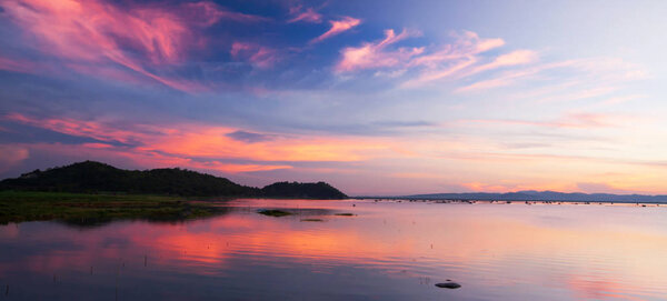 Beautiful twilight sky above a tropical lake, gently light pink clouds against the blue sky at dusk, soft reflection of clouds and mountains on surface of freshwater, rural scene of Suphan Buri, Thailand.