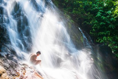 Yazın tropikal şelalenin tatlı sularında oynayan mutlu, çıplak göğüslü Asyalı çocuk, şelale serpintilerinin şeffaf sanatı. Khao Lak, Tayland. Uzun pozlama. Hareket bulanıklığı.