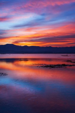 Gölün üzerinde dramatik bulutlar ve günbatımı gökyüzü ve arka planda dağ, renkli bulutlar ve su yüzeyine yansıyan gökyüzü, gölde balıkçı teknesinde balıkçı. Phayao Gölü, Tayland.