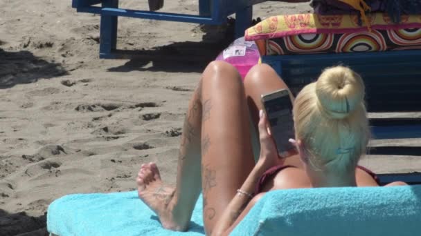 一个带着手机和日光浴在海滩上打字的金发女孩 — 图库视频影像