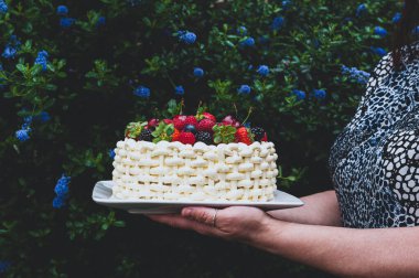 Çiçekli arka planda böğürtlenlerle süslenmiş güzel bir yaz pastası taşıyan kadın..