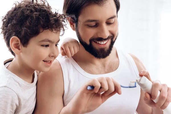 Close up. Bearded father shows young son how to squeeze toothpaste on toothbrush. — Stock Photo, Image