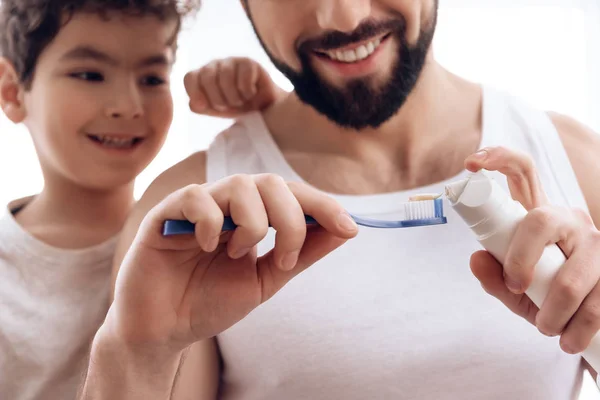Nahaufnahme. bärtiger Vater zeigt kleinem Sohn, wie man Zahnpasta auf Zahnbürste drückt. — Stockfoto