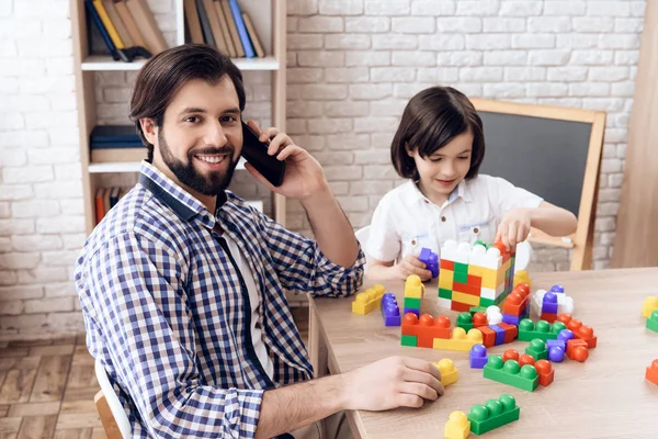 Joyeux père parle au téléphone tandis que le fils joue avec le constructeur de jouets . — Photo