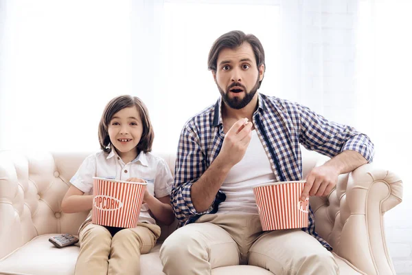 Bearded father with small son is watching thrilling film, eating popcorn.