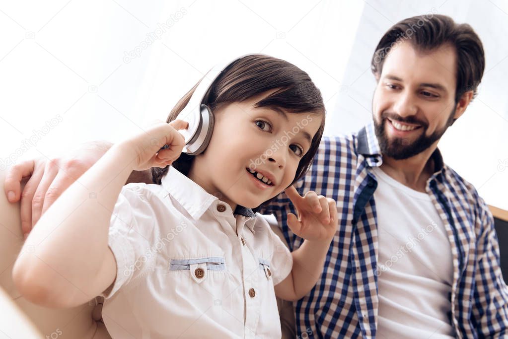 Teenage boy in headphones listens to music, sitting with happy father on couch.