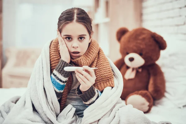 Menina com frio no lenço e cobertor com termômetro nas mãos em casa . — Fotografia de Stock