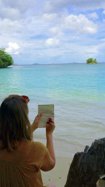Blonde caucasian woman on the beach drawing on notebook with sea tropical landscape.