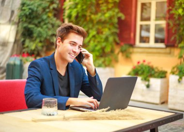 Cep telefonu olan yakışıklı genç bir iş adamı bir yaz kafesi avlusunda dizüstü bilgisayarda çalışıyor.