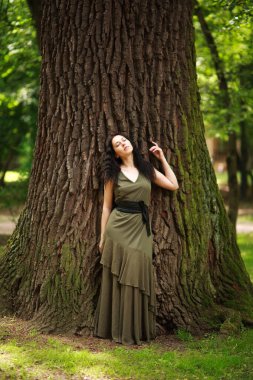 Yeşil elbiseli genç kadın, bir orman parkındaki büyük bir ağacın yanında meditasyonla rahatlıyor. Doğanın saflığı ve çevreyle bütünlük kavramı.