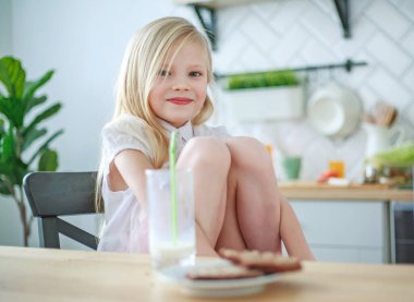 Küçük kız bir bardak süt ve kurabiyeyle masada oturuyor, evde sağlıklı bir kahvaltı.