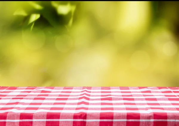 Una mesa vacía con un mantel a cuadros rojo y un fondo bokeh abstracto en la mañana en el jardín. Útil para montaje en exhibición de alimentos y productos . —  Fotos de Stock