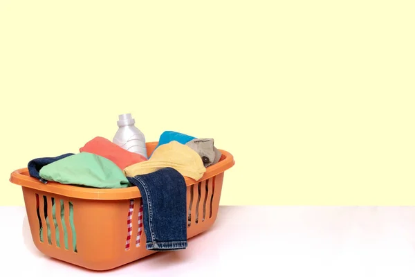 Primer plano de una cesta de lavado naranja con ropa sucia de colores y una botella de detergente blanco sobre una mesa brillante sobre fondo amarillo claro. Espacio para el montaje del producto . —  Fotos de Stock