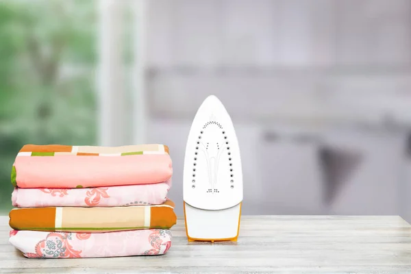 Household laundry ironing. Close-up of a yellow electrical iron and a stack of ironed clothes on white board over blurred curtains background. Advertising laundry service.