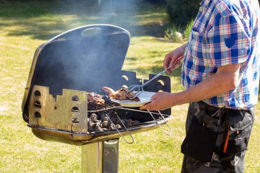 Kömür lokantasında bir adam biftek, ızgara sosis ve tavuk kanadı pişiriyor ve barbekü maşası kullanıyor. Güneşli bir bahar gününde yeşil çimenli arka planda ızgaraya odaklan. Mangal konsepti. Makro.