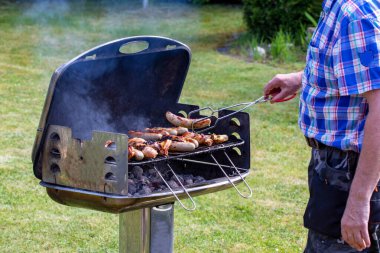 Kömür lokantasında bir adam biftek, ızgara sosis ve tavuk kanadı pişiriyor ve barbekü maşası kullanıyor. Güneşli bir bahar gününde yeşil çimenli arka planda ızgaraya odaklan. Mangal konsepti. Makro.