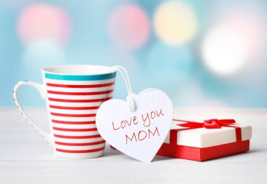 Anneler günü tebrik kartı. Hediye ya da hediye kutusu olan bir kupanın dekoratif kompozisyonu ve üzerinde yazılar olan kalp şeklinde bir etiket. Anneni parlak bir masanın üzerinde bulanık bir arka plan üzerinde seviyorum..